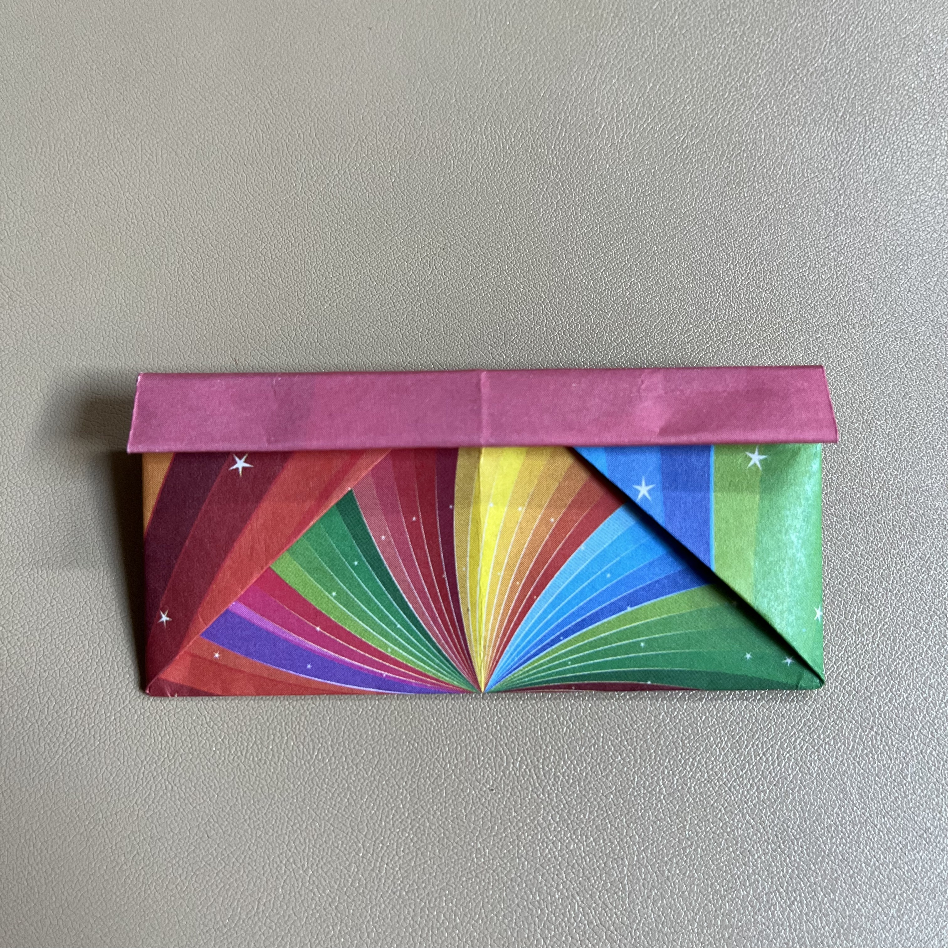 A colorful origami envelope with a rainbow pattern and stars on the lower section and a solid pink top flap, set against a plain background.