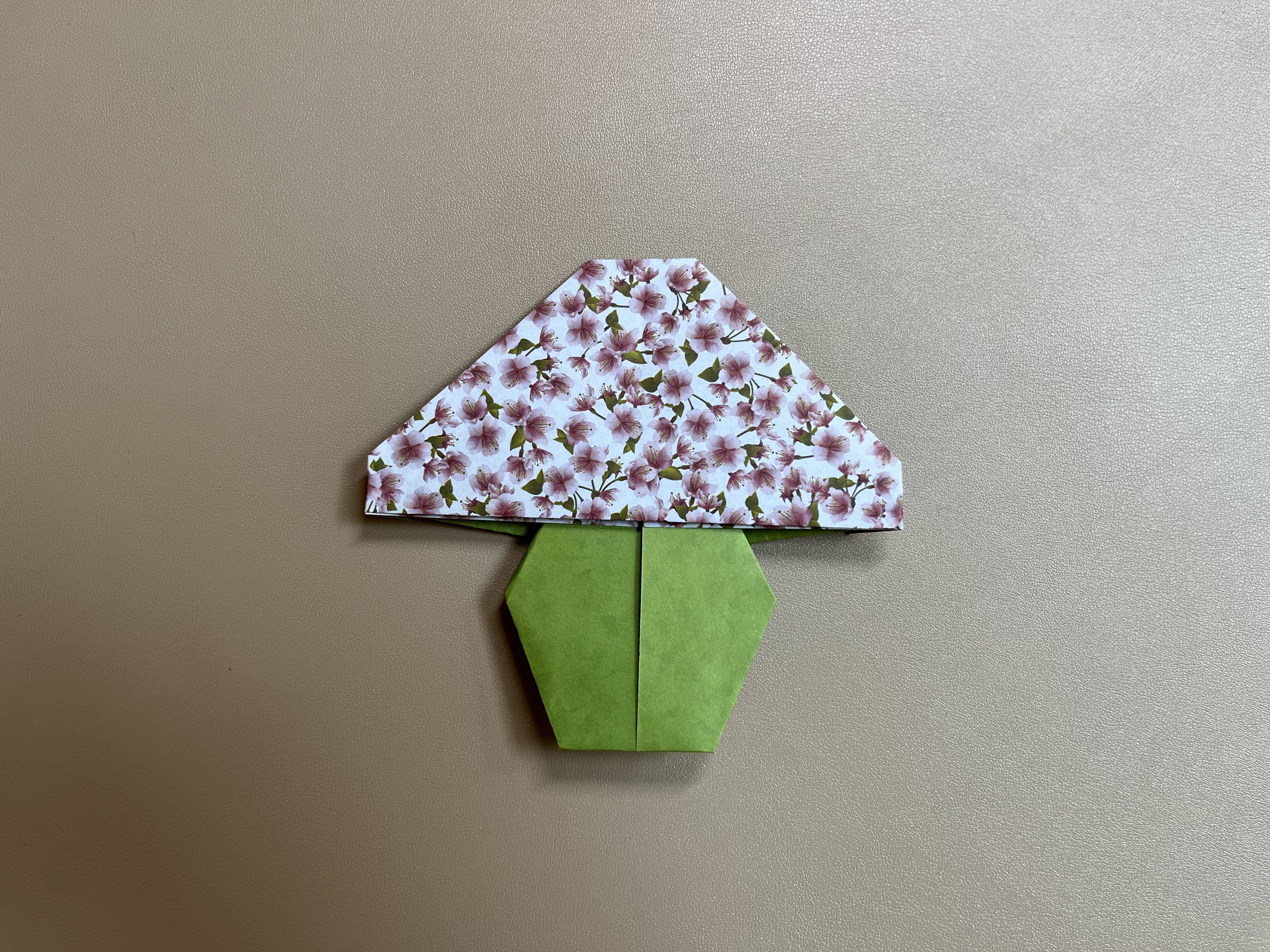 An origami mushroom made from decorated paper rests on a light brown background. The mushroom's cap is white with a detailed floral pattern in pink and green, while its stem is crafted from plain green paper folded to form two hexagonal shapes.
