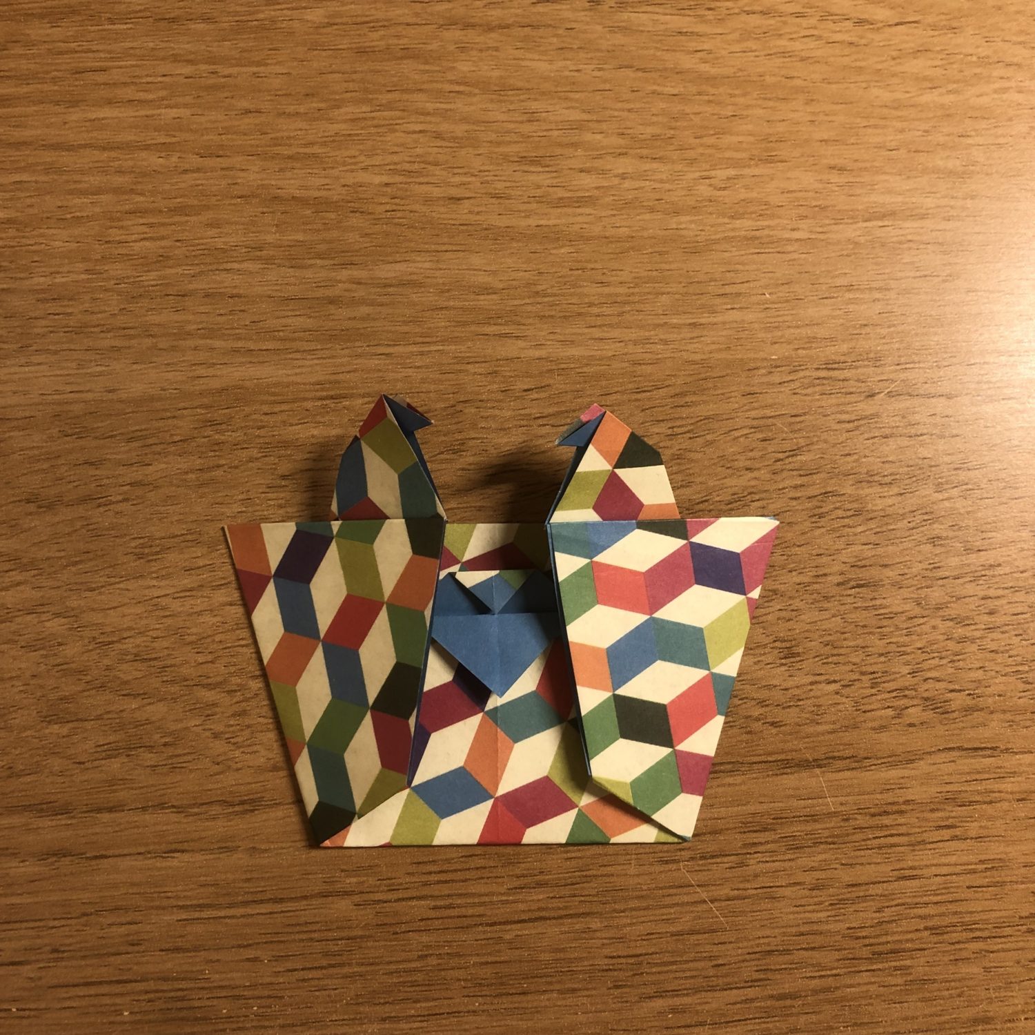 A colorful, geometric-patterned origami piece sits on a wooden surface. The artwork features two crane-like figures side by side with their beaks pointing upward, crafted from paper with a vibrant pattern of interconnected hexagons.