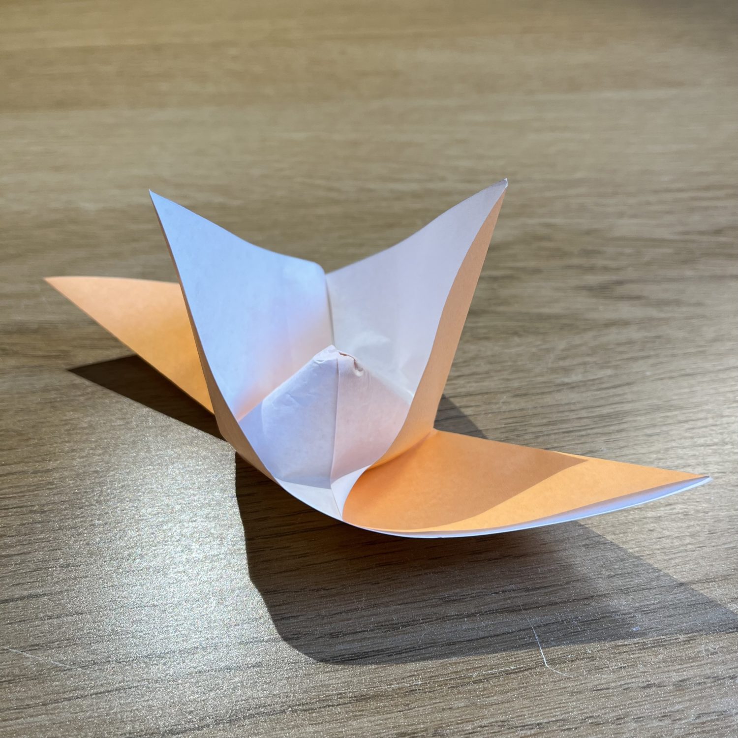A partially folded origami project made from a single sheet of orange and white paper, placed on a wooden surface. The paper appears to be in the process of forming a flower or boat-like shape.