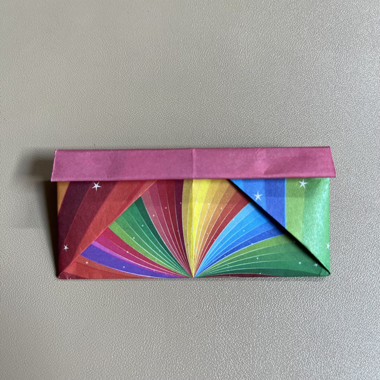 A colorful origami envelope with a rainbow pattern and stars on the lower section and a solid pink top flap, set against a plain background.