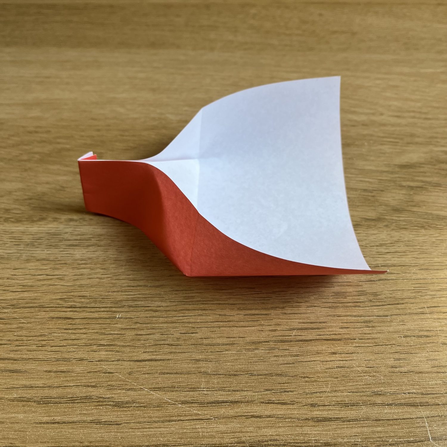 An origami dustpan  featuring a red and white paper model, partially folded with one side lifted, sits on a wood surface.