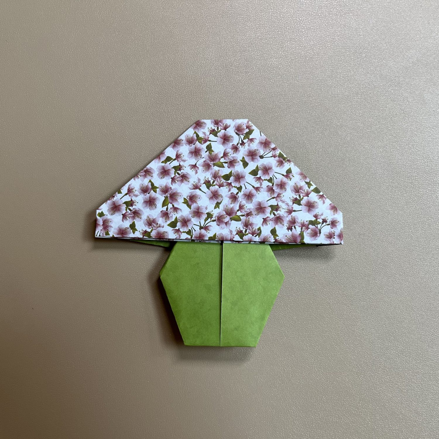 An origami mushroom made from decorated paper rests on a light brown background. The mushroom's cap is white with a detailed floral pattern in pink and green, while its stem is crafted from plain green paper folded to form two hexagonal shapes.