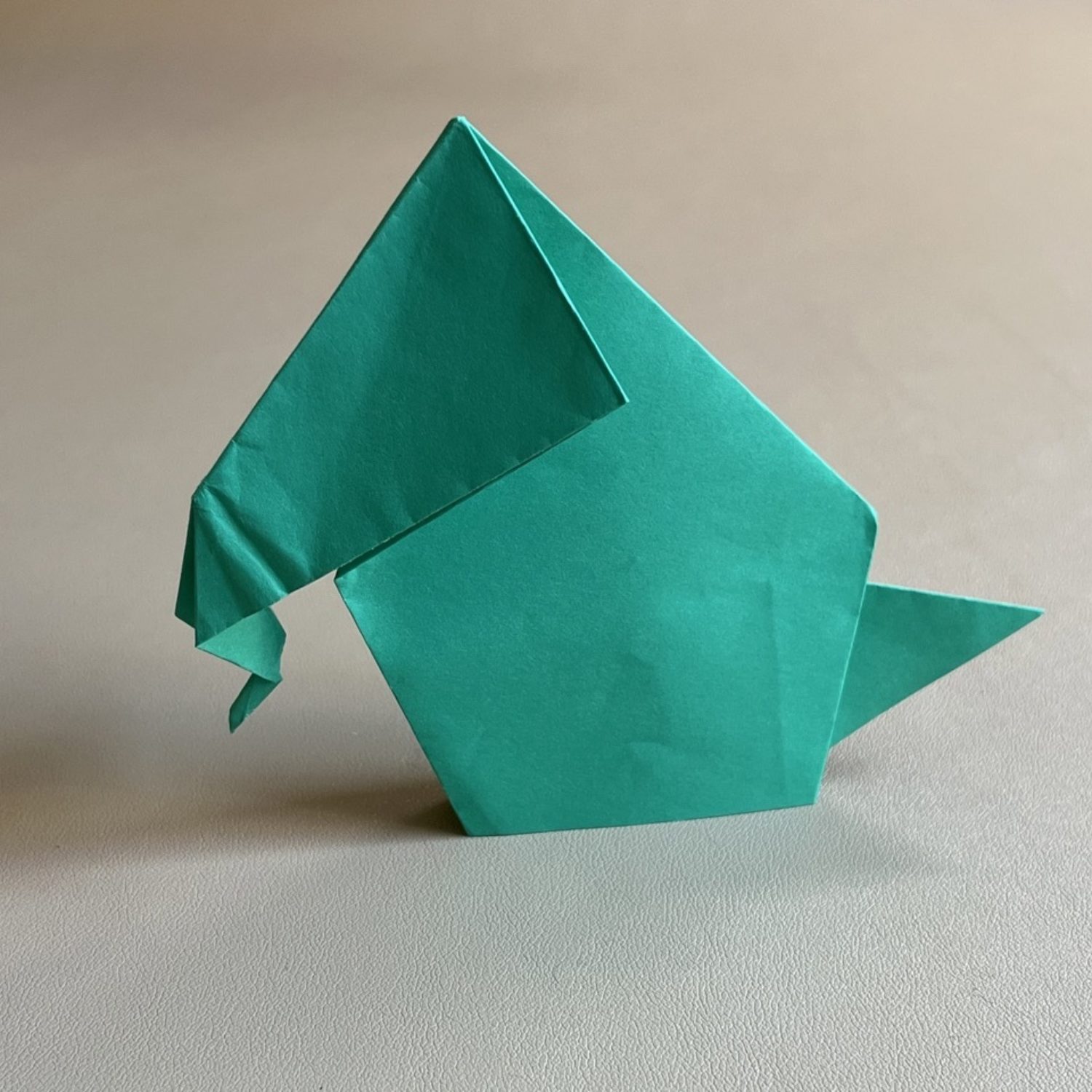 A green origami elephant, neatly folded from a single sheet of paper, sits on a smooth, light-gray surface. Its trunk is curved downward, and the figure captures the basic features of an elephant. The background is plain and unobtrusive.