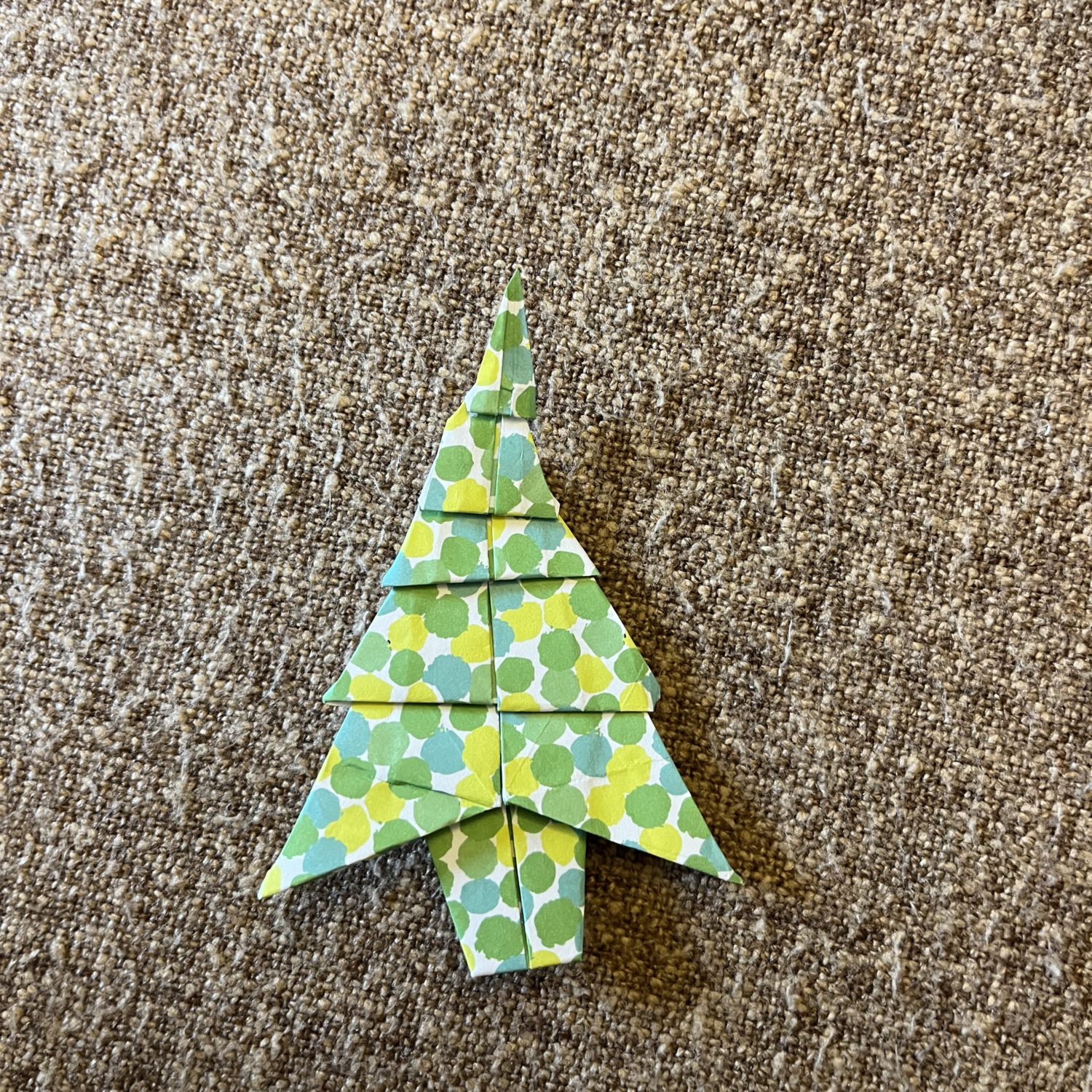 A handmade origami Christmas tree made from green and white polka-dotted paper is lying on a textured, brown fabric surface.