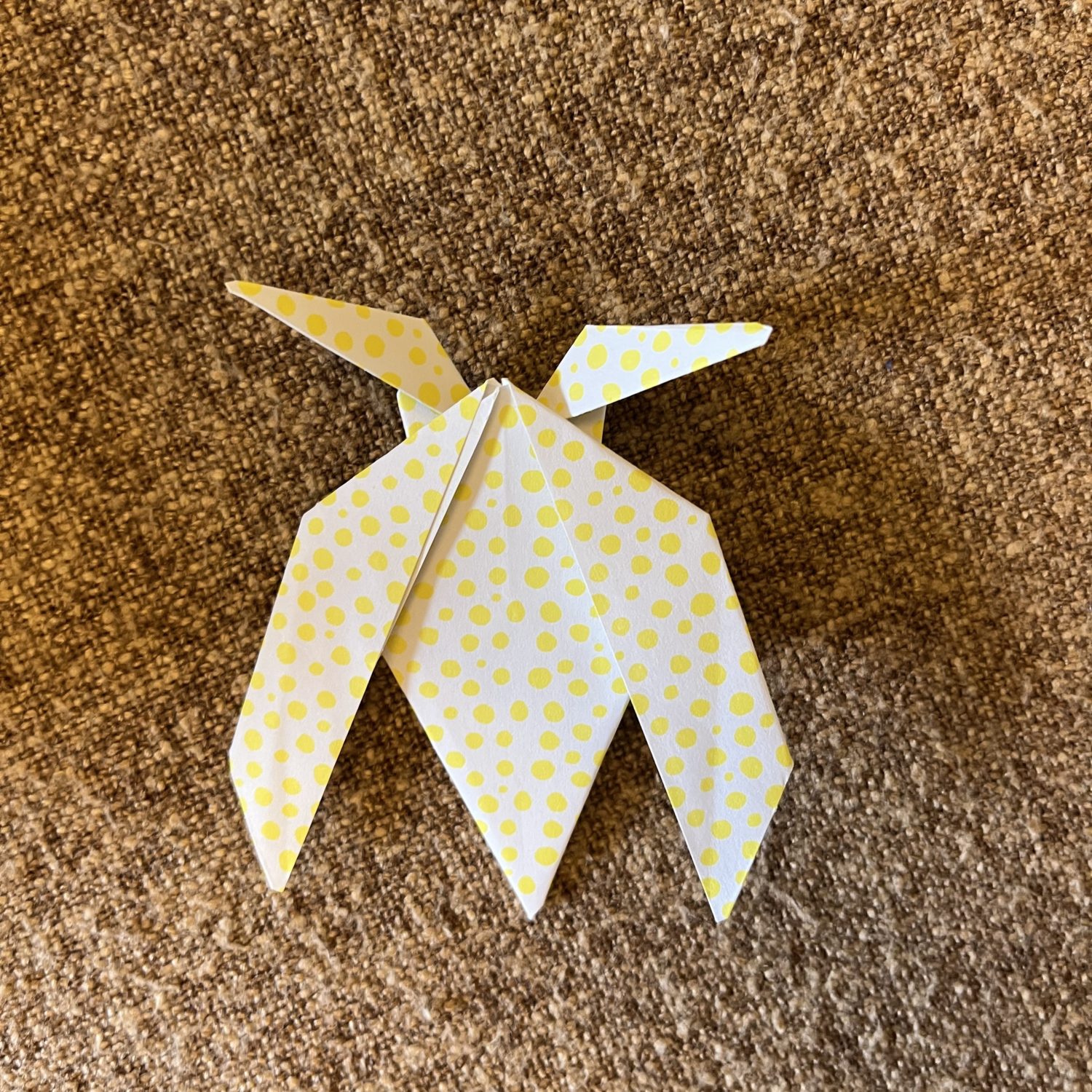 A yellow and white origami moth with a polka dot pattern is placed on a textured brown surface. The moth has intricately folded wings and pointed details, showcasing the art of paper folding.