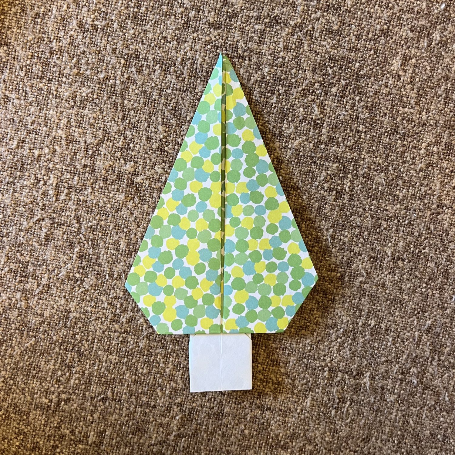 A paper origami tree with a geometric, polka-dot pattern in shades of green, blue, yellow, and white sits on a textured brown surface. The tree has a white rectangular base, resembling a tree trunk.