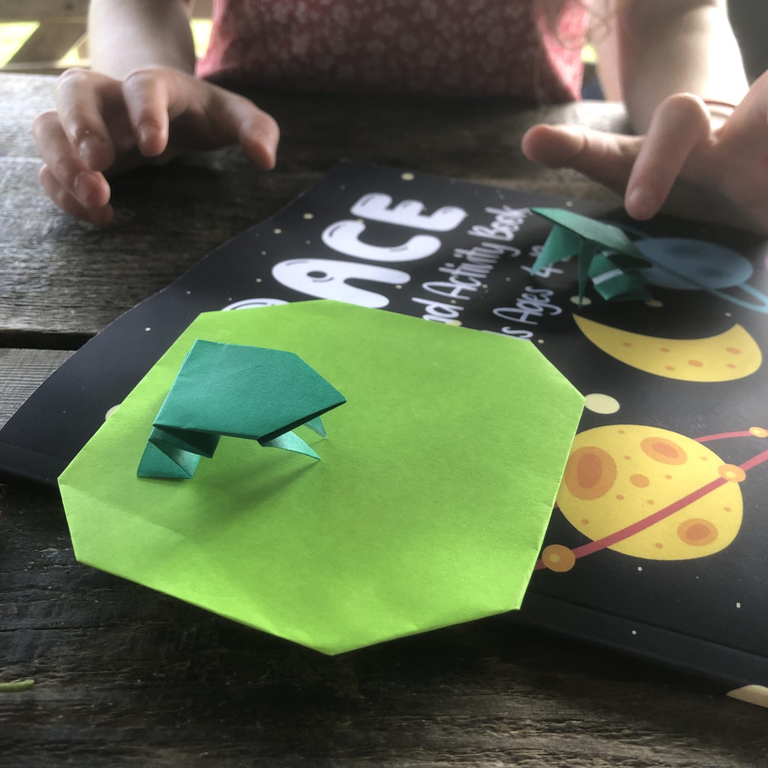 A child with origami paper frogs on a green paper lily pad and a space-themed activity book. The child’s hands are visible, reaching toward the paper frogs. The scene is set on a wooden surface, with the child wearing a patterned top.