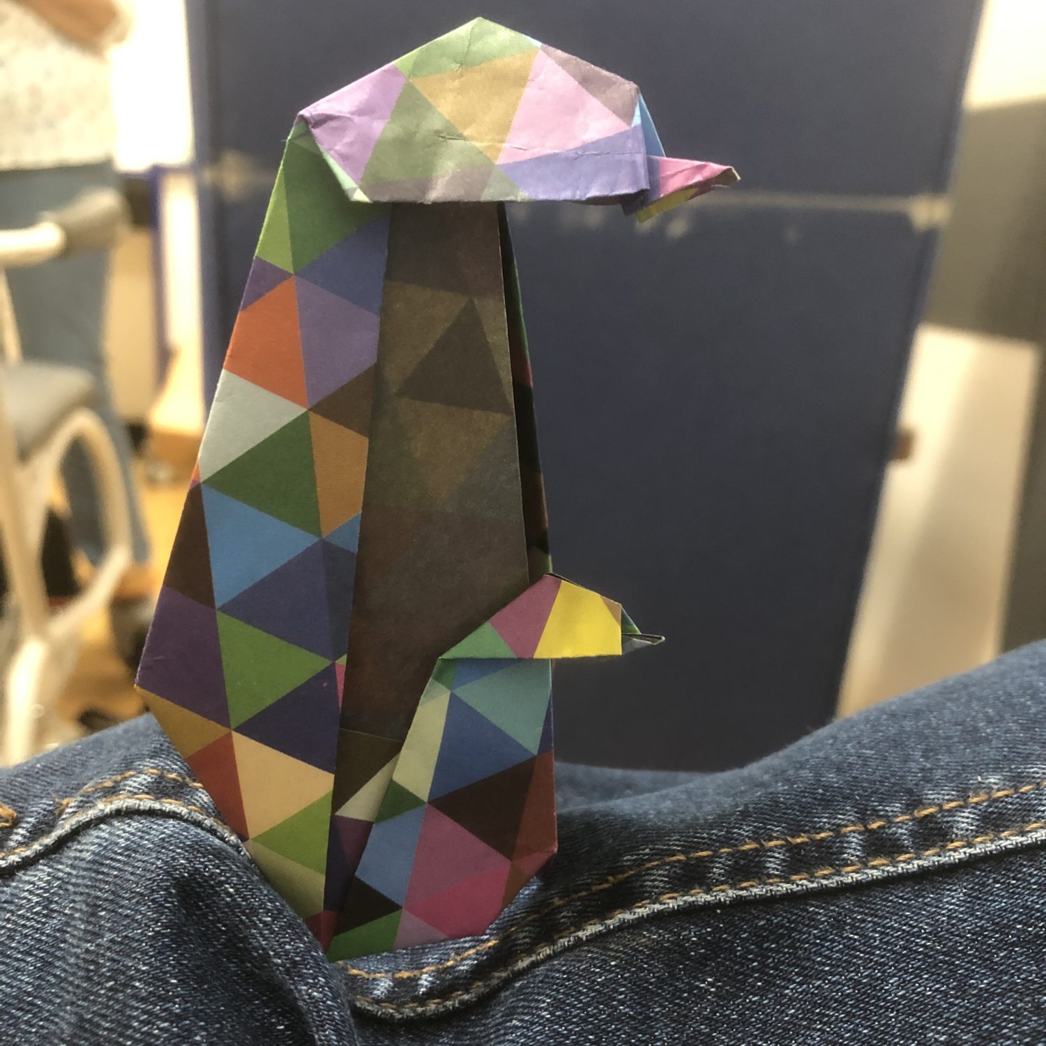 A colorful, geometric paper origami penguin perched on a person's denim-covered knee. The origami features a mix of vibrant colors and patterns.