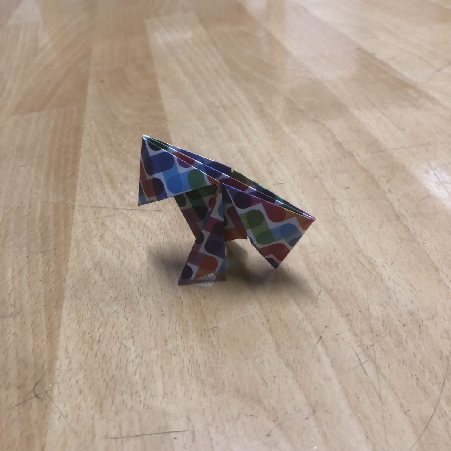 A small, colorful origami dinosaur is placed on a wooden surface. The dinosaur is crafted from paper with a multicolored pattern consisting of various shapes. The surface has a light wood grain finish.