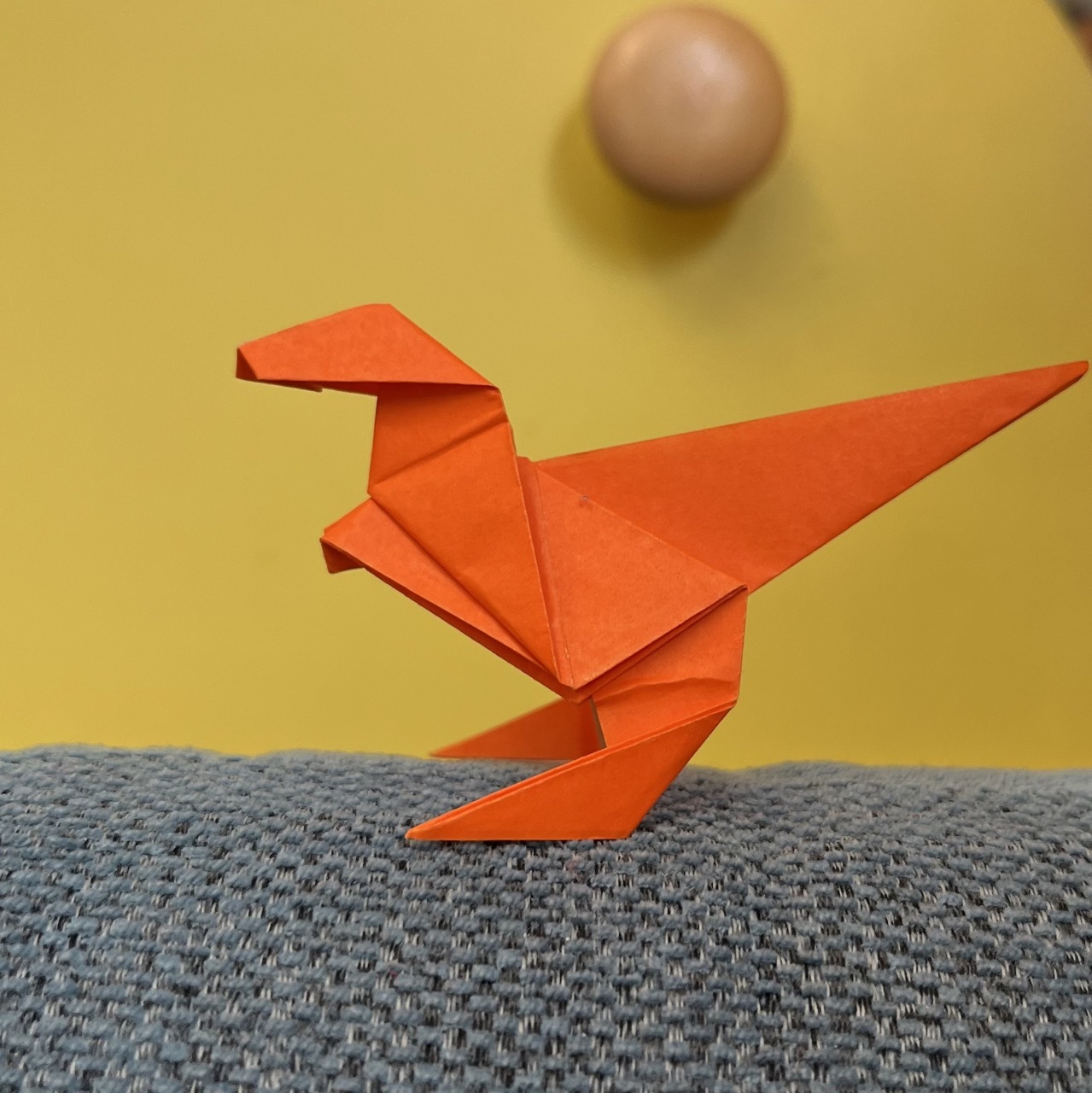 An orange origami dinosaur perched on a grey fabric surface with a blurred beige ball in the background.