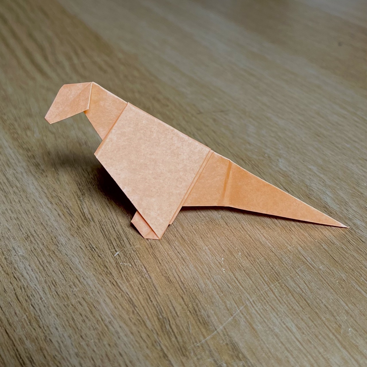 A origami origami dinosaur, crafted from a single piece of paper, displayed on a wooden surface. the dinosaur has a prominent head and tail, standing with a slanted posture.
