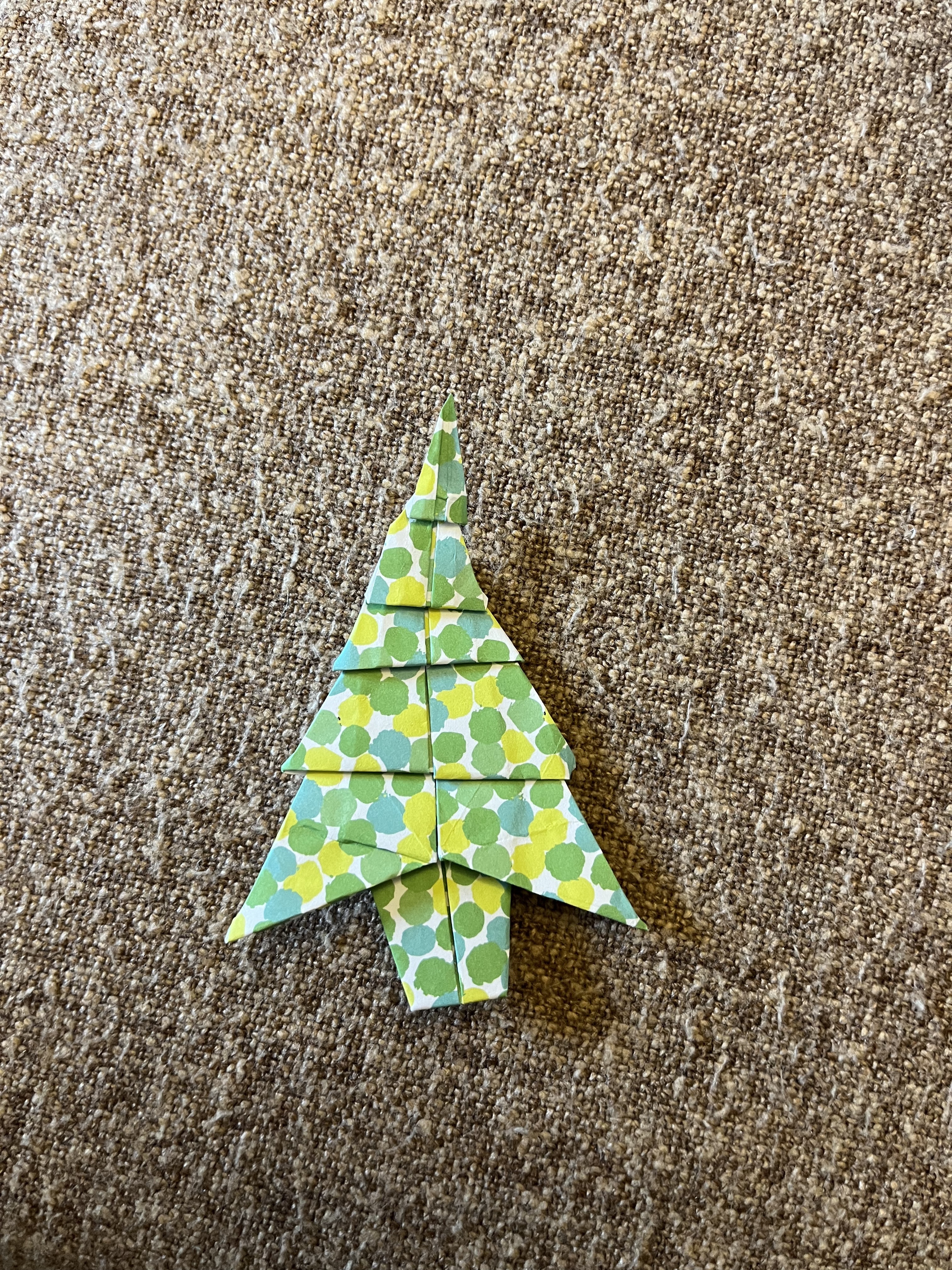 A handmade origami Christmas tree made from green and white polka-dotted paper is lying on a textured, brown fabric surface.