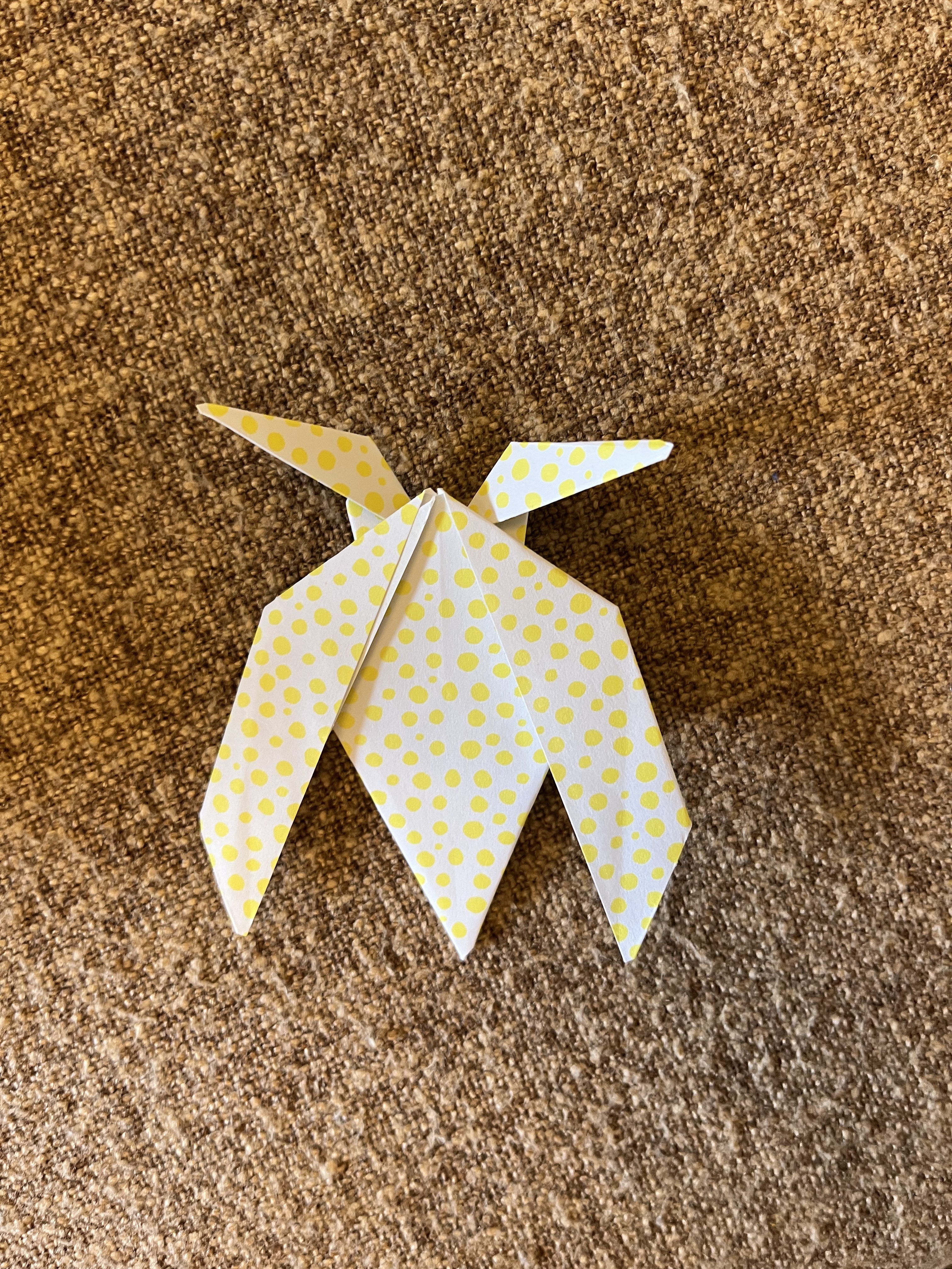 A yellow and white origami moth with a polka dot pattern is placed on a textured brown surface. The moth has intricately folded wings and pointed details, showcasing the art of paper folding.