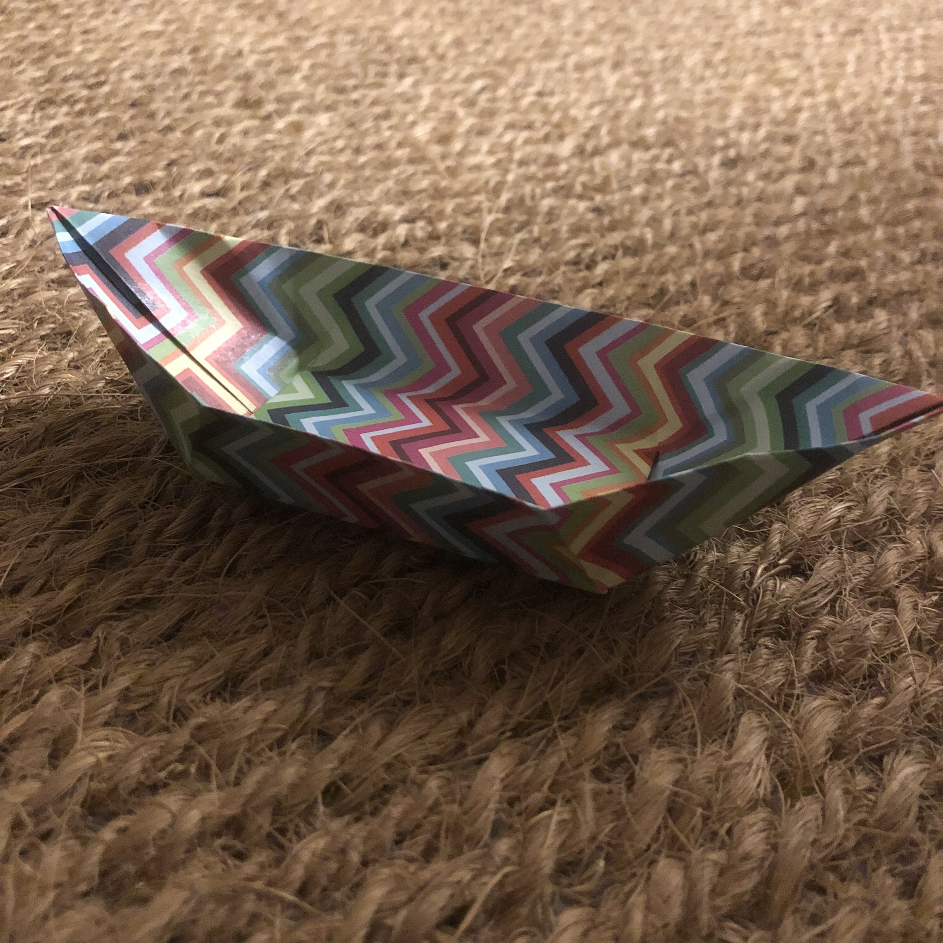 A colorful paper boat with a zigzag pattern in various shades of blue, red, pink, green, and yellow rests on a textured beige fabric surface. The boat is carefully folded, showing crisp edges and angles.