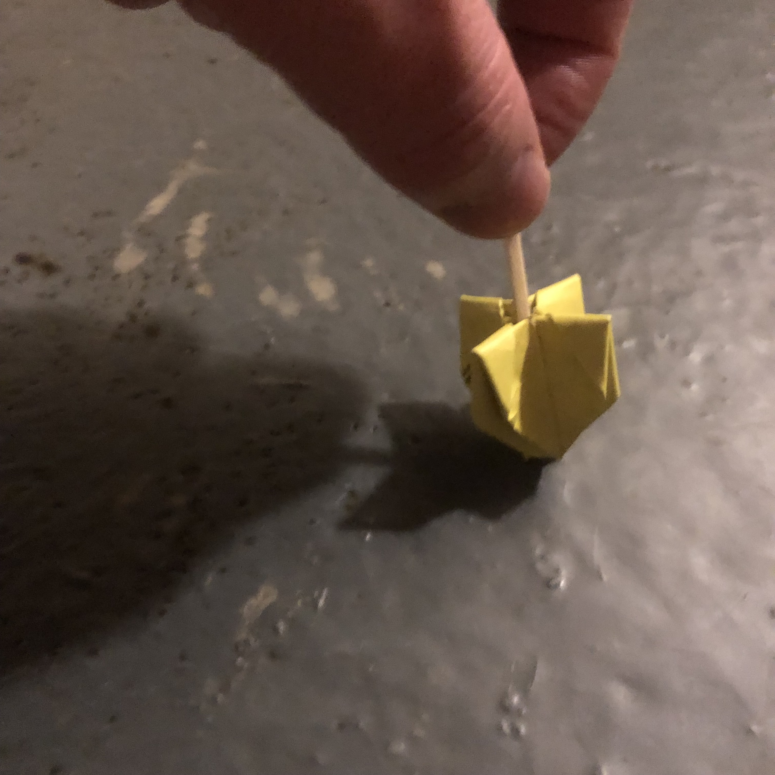 A hand holding a small yellow origami Dreidel with the help of a toothpick, shown against a textured gray background, casting a shadow.