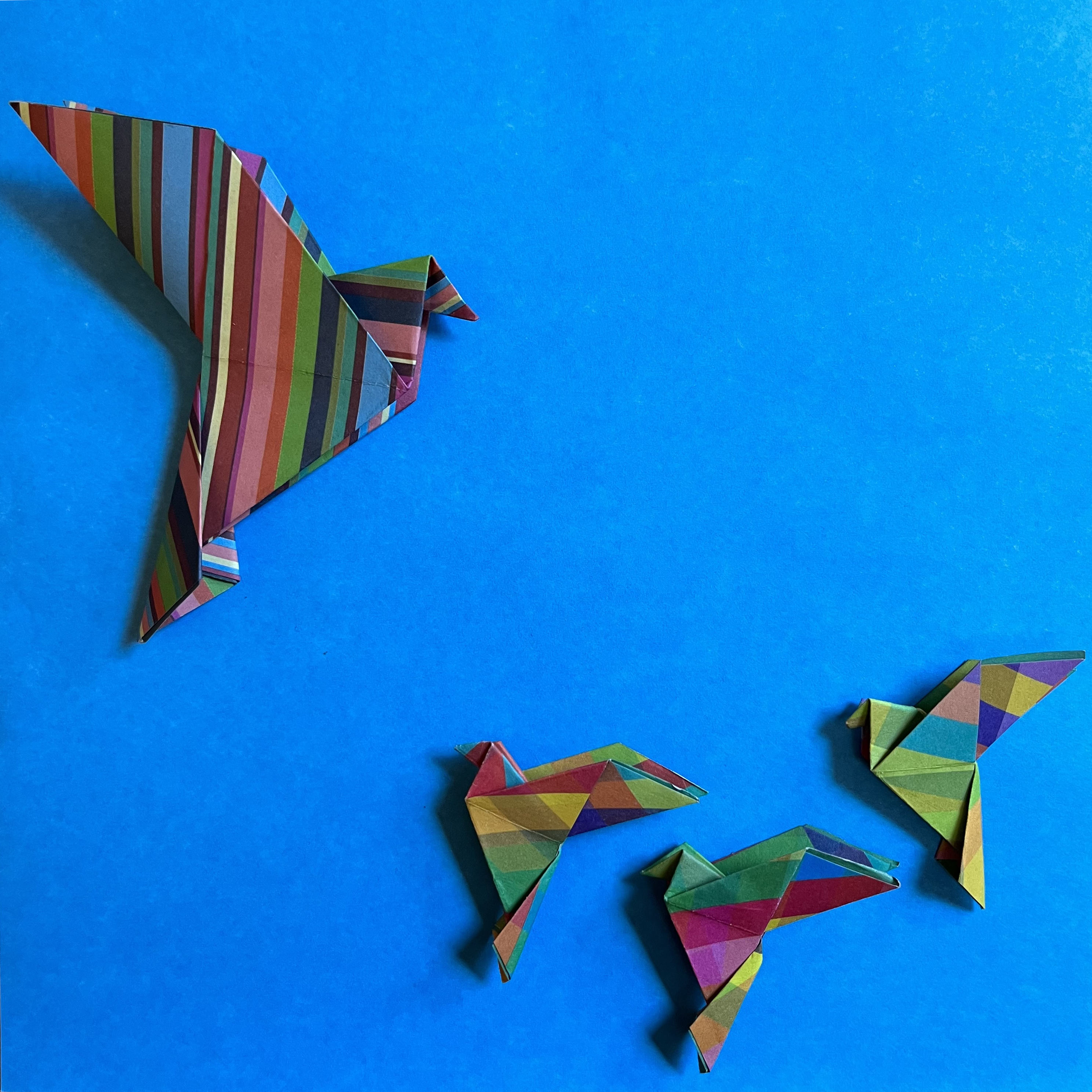 Four multicolored origami birds are arranged on a vibrant blue background. One bird, larger and striped with vertical bands of color, appears to be leading, while the three smaller birds, each with varied geometric patterns, follow closely behind.