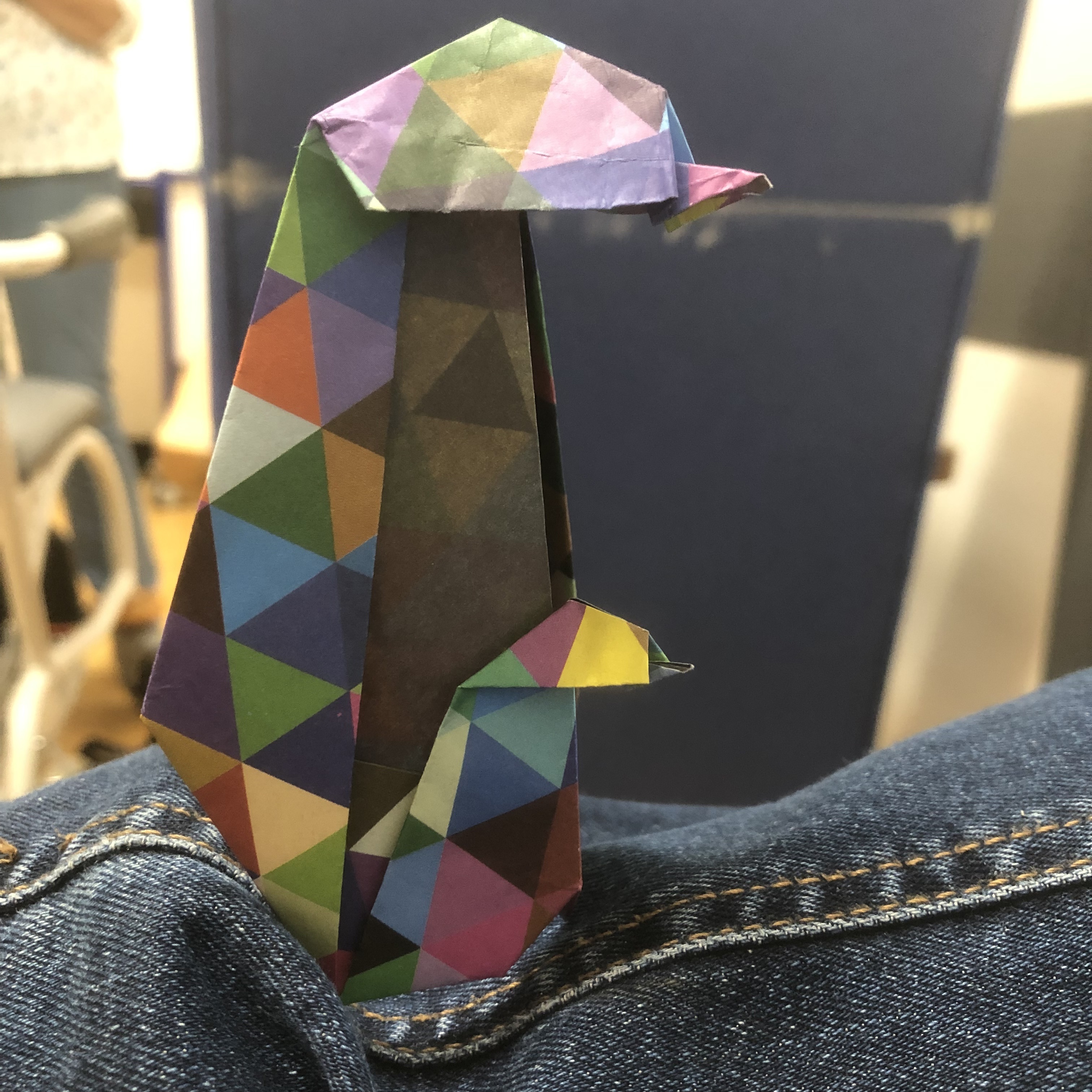 A colorful, geometric paper origami penguin perched on a person's denim-covered knee. The origami features a mix of vibrant colors and patterns.