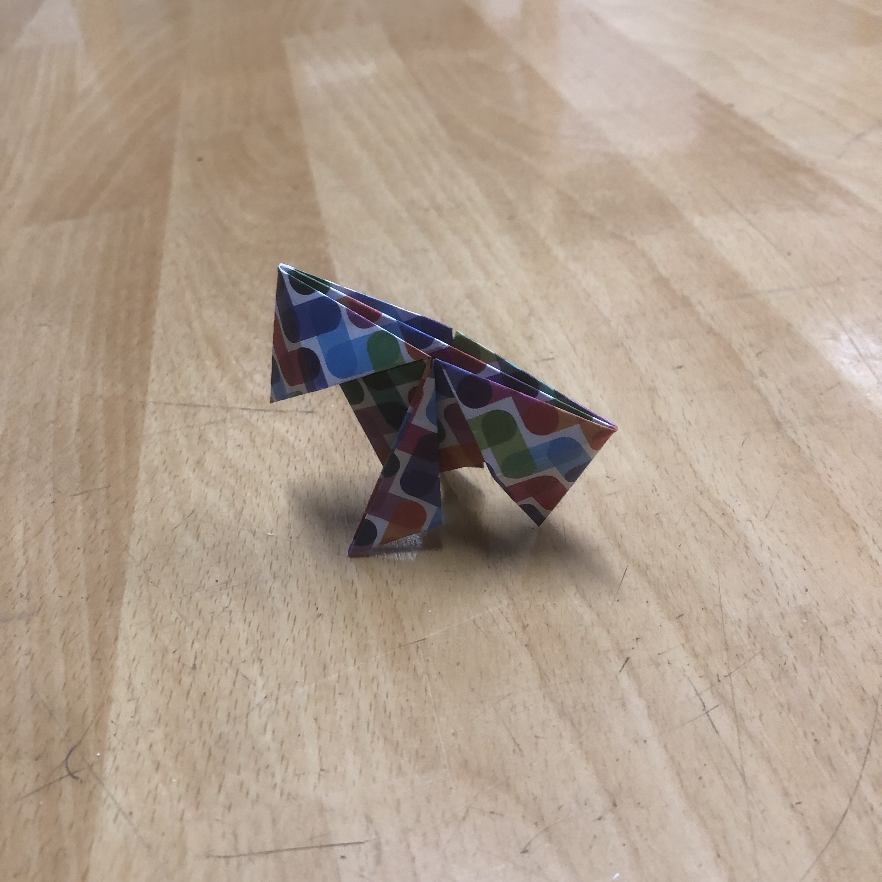 A small, colorful origami dinosaur is placed on a wooden surface. The dinosaur is crafted from paper with a multicolored pattern consisting of various shapes. The surface has a light wood grain finish.