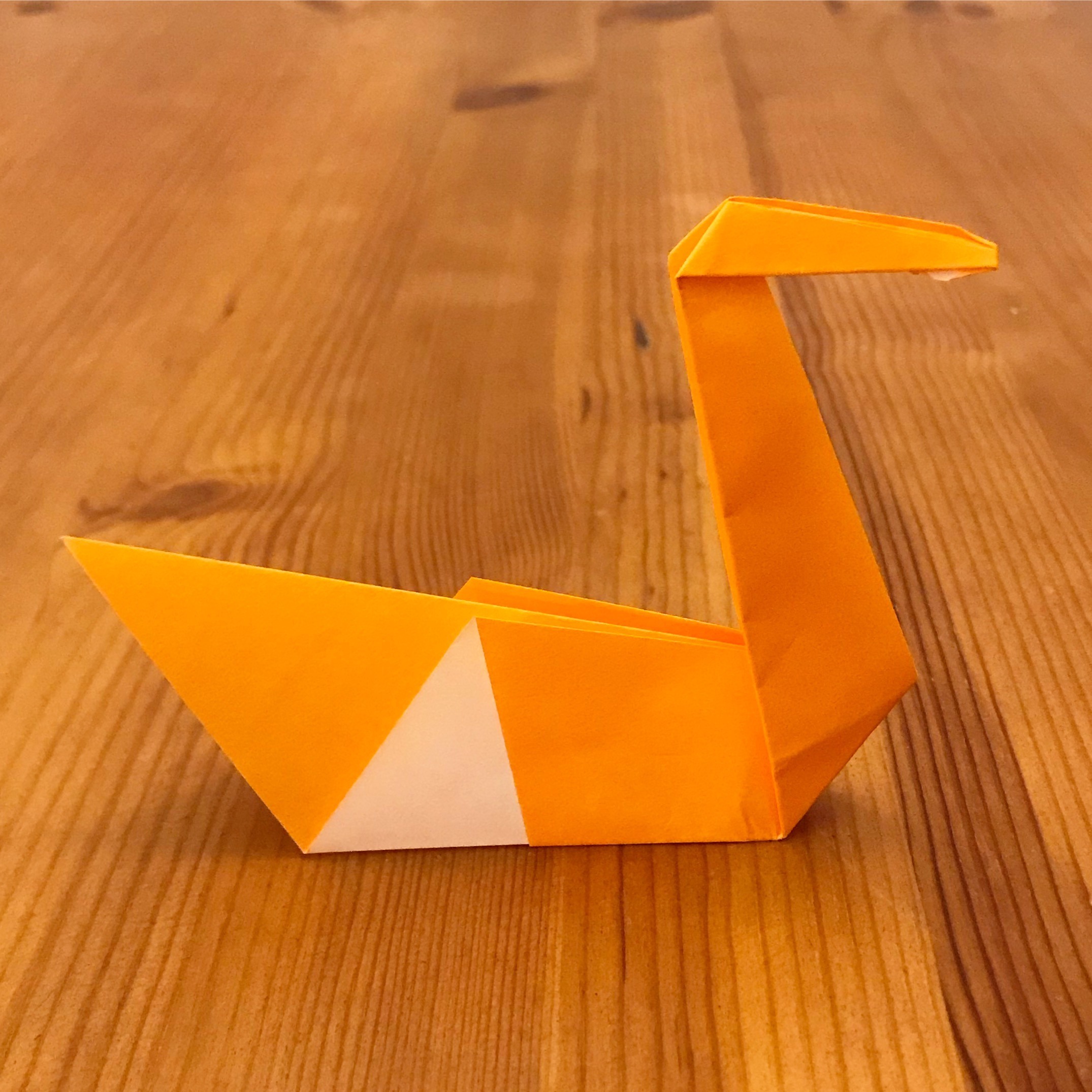 A yellow and white origami swan displayed on a wooden surface, highlighting its elegant neck and triangular wings.