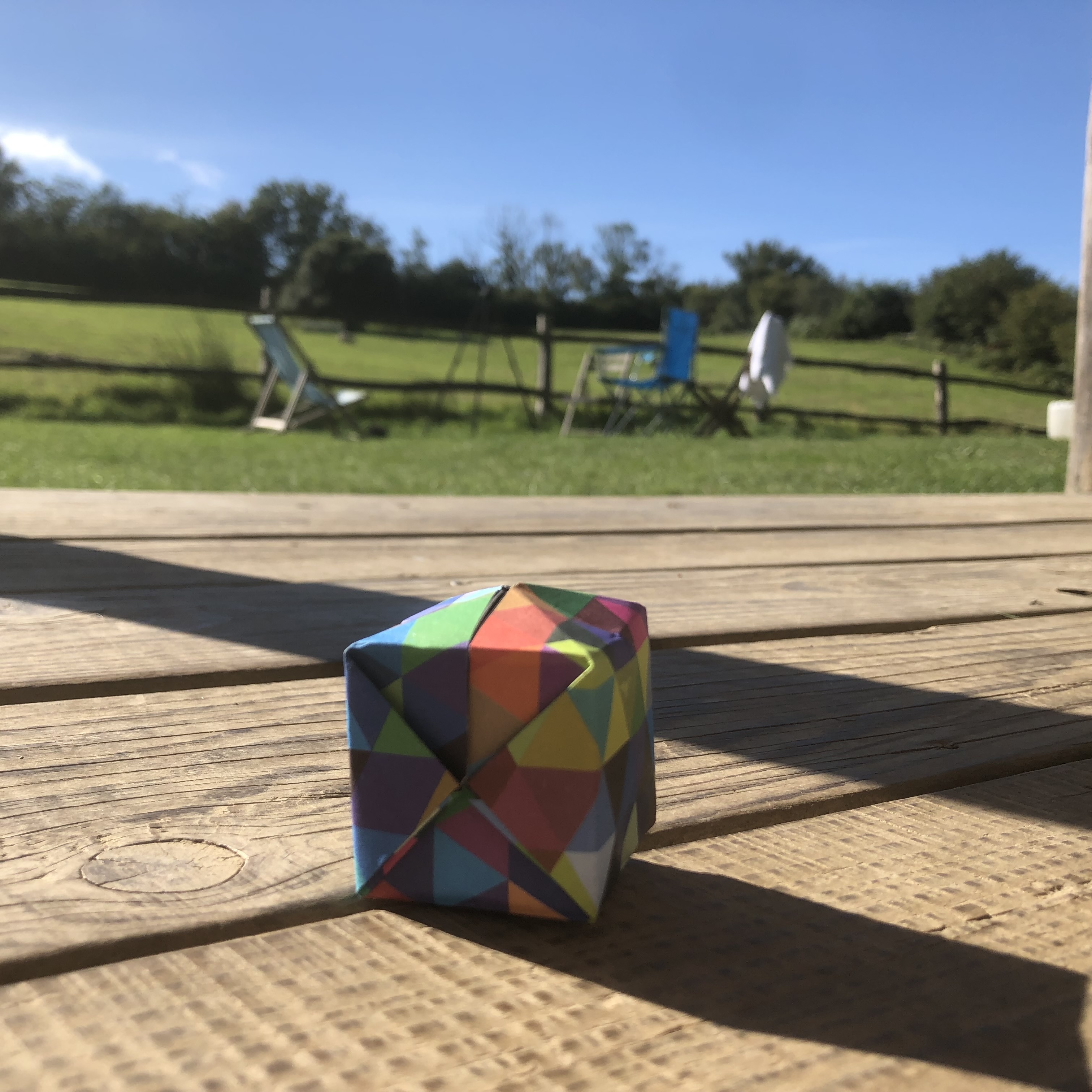 A colorful, geometric-patterned origami cube sits on a wooden deck in the foreground. In the background, a grassy field with trees can be seen, along with some scattered chairs and a blue sky with a few clouds. Sunlight casts shadows on the deck.