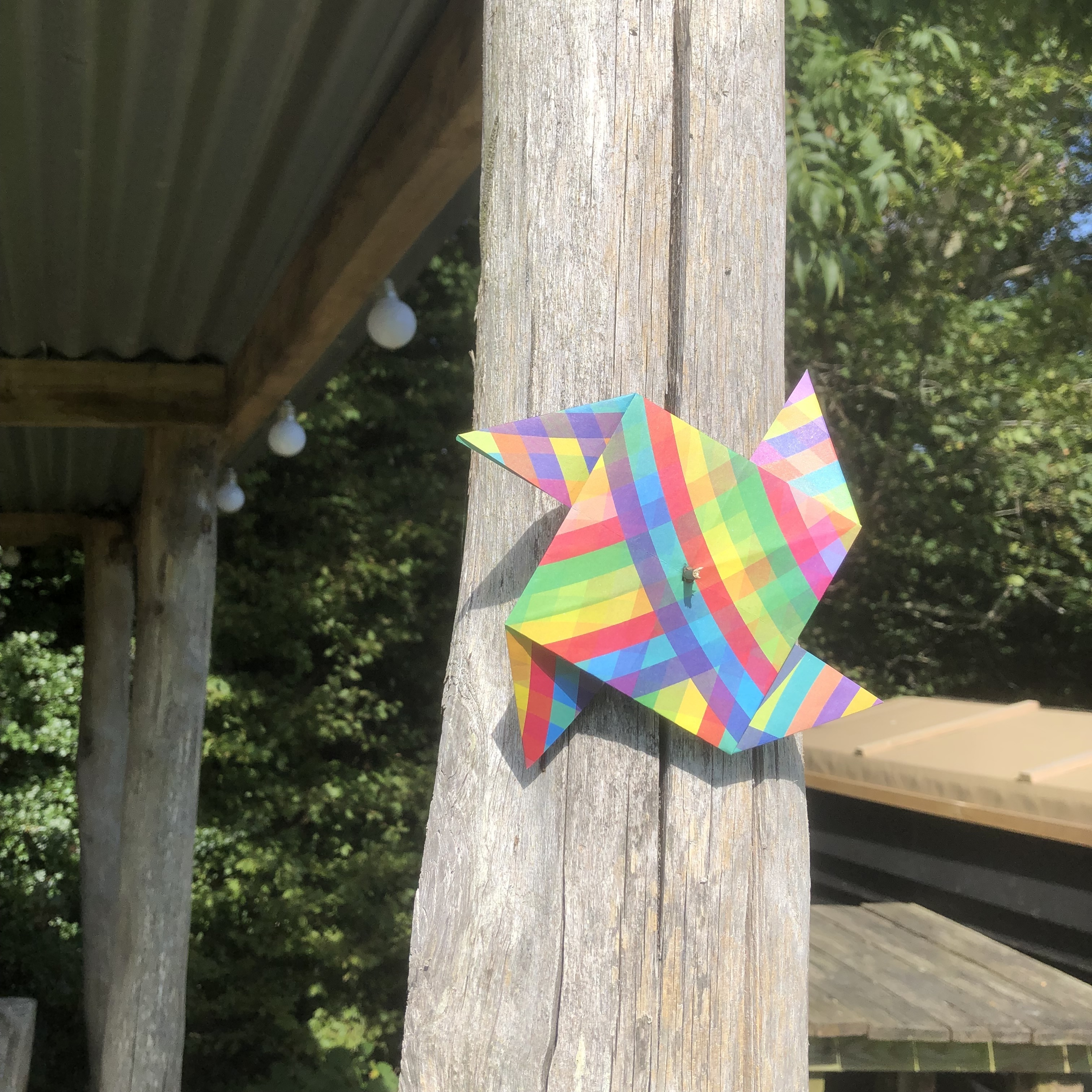 A colorful origami paper pinwheel with a rainbow checkered pattern is pinned to a wooden post under a corrugated metal roof. In the background, there are more wooden posts, a few string lights, and green foliage. The scene appears to be outdoors on a sunny day.